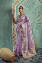 A woman models a lavender purple silk saree with a beige border and gold embroidery, posed against a textured teal wall with decorative arches and a large vase.