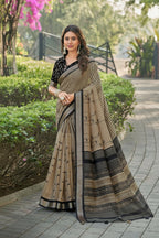 A woman stands outdoors on a paved walkway wearing a beige and black printed saree with a black blouse. The background shows green hedges, trees, and a garden path.