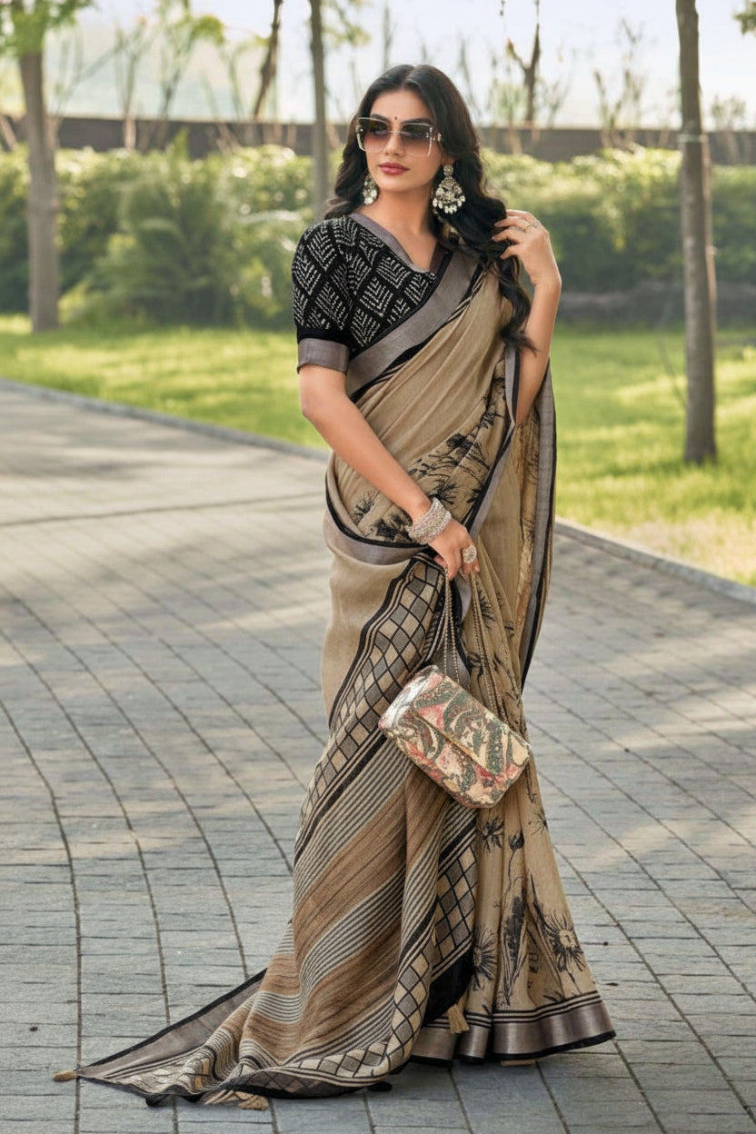 A woman wearing a beige and brown patterned saree with a black printed blouse, standing outdoors on a paved walkway. She is holding a small patterned clutch bag.