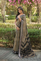 A woman wearing a pleated grey and beige patterned saree stands outdoors in a garden setting with green hedges and pink flowering trees in the background.