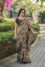 A woman wearing a beige and black printed saree with a black embroidered blouse, standing on a paved pathway in a garden setting.
