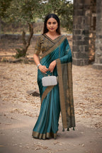 A woman wearing a teal green and gold saree stands outdoors in front of a stone structure. The saree has a gold patterned border and a matching blouse.