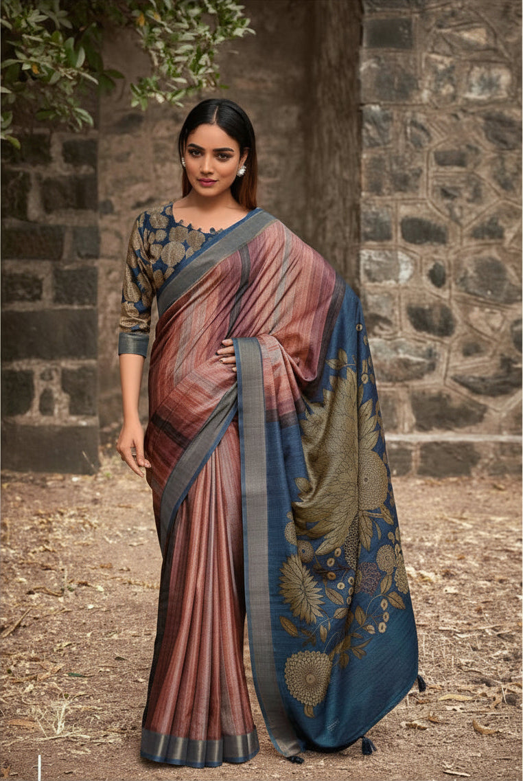 A woman models a traditional Indian saree in an outdoor setting with a stone wall background. The saree has a dusty rose and blue gradient body with a dark blue border and gold floral patterns, paired with a matching patterned blouse.