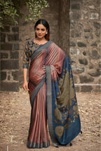 A woman models a traditional Indian saree in an outdoor setting with a stone wall background. The saree has a dusty rose and blue gradient body with a dark blue border and gold floral patterns, paired with a matching patterned blouse.