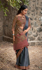 A woman modeling a traditional Indian saree in an outdoor setting with a stone wall and tree branches in the background. The saree has a brown and gold patterned blouse, a rust-colored border, and a dark gray-blue skirt with a brown border.