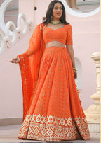 A woman modeling an orange embroidered lehenga choli set with a matching dupatta, featuring intricate gold and white geometric patterns on the skirt and border, set against a light pink architectural background.