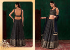 A woman modeling a black and gold embellished lehenga choli set, shown in two views: a back view on the left and a front view on the right, set against a dark green studio backdrop with a patterned rug.